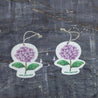 A top-view comparison showing the Purple hydrangea pop-up sponge in dry versus wet states. On the right : the sponge appears flat and compact in its dry form. On the left: it has expanded into a thicker cleaning pad after hydration. Both clearly display the charming Purple hydrangea pattern and feature an attached hanging string. Photographed against a dark grey textured background, this eco-friendly kitchen cleaner demonstrates remarkable size expansion when water is absorbed.