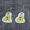 Top-view comparison of the holiday dinosaur pop-up sponge in dry versus wet states. The right side shows the compact, flat disc in its dry form, while the left demonstrates the expanded, thicker cleaning pad after water absorption. Both feature an adorable holiday dinosaur wearing a red Santa hat with matching bowtie, complete with practical hanging cord. Shot against a dark grey textured background.