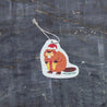 Top view of an eco-friendly cellulose pop-up kitchen sponge with hanging cord, featuring an adorable holiday beaver design wearing a red Santa hat and holding a red gift. This sustainable cleaning tool transforms from completely flat when dry to fully expanded when wet, combining festive cheer with practical functionality. Made from biodegradable plant-based cellulose, the seasonal sponge adds holiday spirit to your kitchen routine while being environmentally responsible.