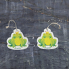This top-view comparison showcases the Frog Prince pop-up sponge in both dry and wet states. The right image displays the sponge in its dry form - flat and compact, while the left demonstrates its expanded, thicker cleaning pad state after hydration. Both versions clearly feature the charming Frog Prince design and come with an attached hanging string. Photographed against a dark grey textured background, this eco-friendly kitchen cleaner exhibits remarkable size expansion when water is absorbed