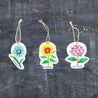 Top view of three eco-friendly pop-up sponges featuring beautiful floral designs. The set includes: 1) Sunflower, 2) Purple hydrangea, and 3) Blue daisy patterns. These circular kitchen sponges are displayed on a dark grey textured background. Made from biodegradable cellulose with attached strings, they combine practical cleaning with the beauty of nature in your everyday routine.