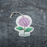 Top view of an eco-friendly cellulose pop-up kitchen sponge with hanging cord, featuring a lovely purple hydrangea design. This sustainable cleaning tool lies completely flat when dry for compact storage, then expands significantly when wet for optimal absorbency. Crafted from biodegradable plant-based cellulose, the sponge showcases a beautiful floral pattern that adds elegance to your kitchen while being gentle on the environment. 