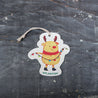 Top view of an eco-friendly cellulose pop-up kitchen sponge with hanging cord, featuring a festive holiday moose wearing a red scarf and holding string lights - compact when dry and expanding when wet for cheerful cleaning.