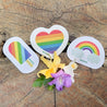 A vibrant display of LGBTQ+ Pride rainbow sponges paired with beautiful hibiscus flowers on a rustic wooden log outdoors. The colorful sponges, each representing a stripe of the LGBTQ+ rainbow flag, symbolize inclusivity, equality, and celebration. They add a joyful and uplifting touch to the scene, complemented by the vibrant hibiscus flowers blooming nearby. The natural setting of the wooden log outdoors creates a harmonious backdrop, highlighting the connection between nature, diversity, and pride.