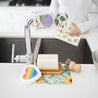 Reusable rainbow heart pop-up sponge with sisal and palm pot scrubber, reusable dishcloths, and a solid dish soap brick placed on top of our natural bamboo dual-layer soap dish.